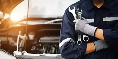 Auto mechanic in uniform holding wrenches stands beside an open car hood in a garage.