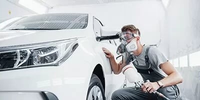 A technician wearing a mask spray-paints a white car in a professional auto body shop.