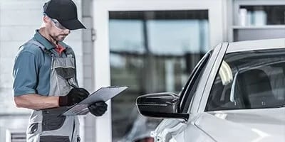 Auto mechanic in uniform reviewing a checklist beside a white car.