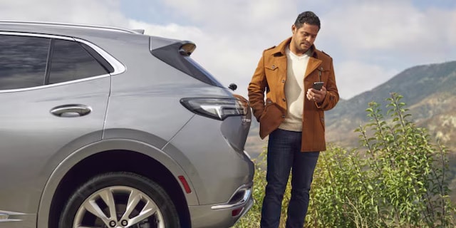 Man in a brown coat stands by a silver SUV, checking his phone with scenic hills in the background.