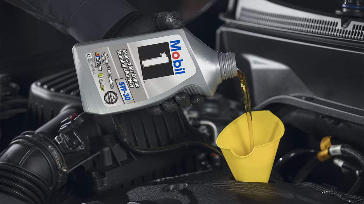 Pouring Mobil 1 5W-30 synthetic motor oil into a yellow funnel under a car hood.