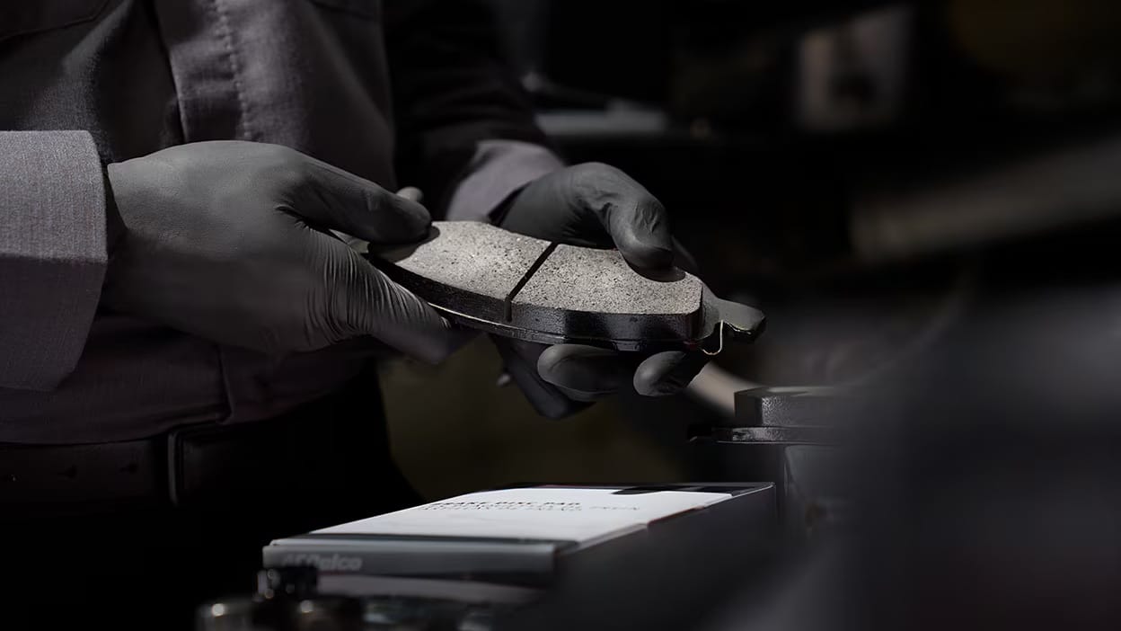 Mechanic wearing gloves holds a car brake pad in a workshop setting.