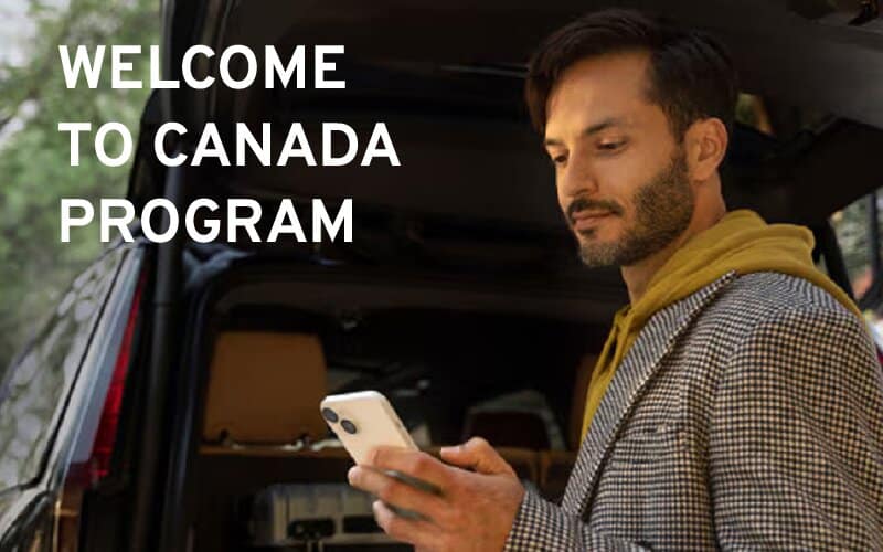 Man checking phone beside car with "Welcome to Canada Program" text. Outdoor setting.