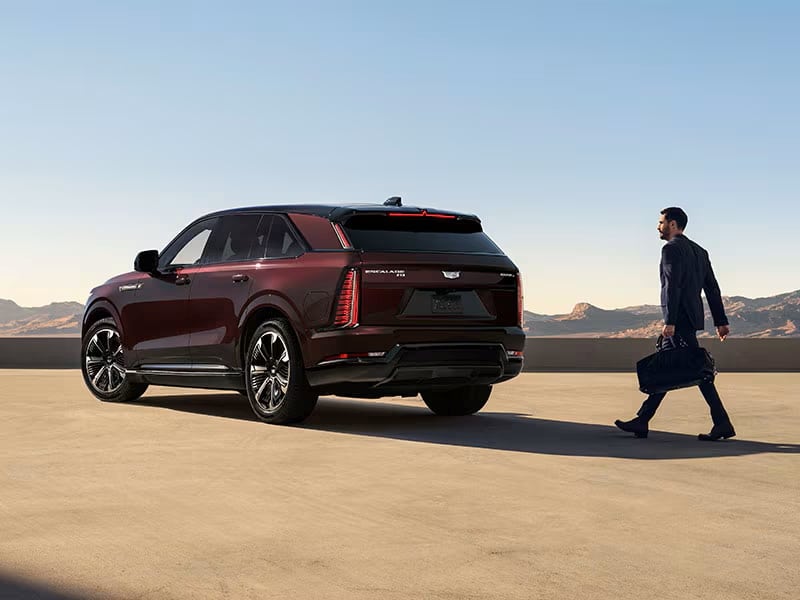 Man walking towards a maroon SUV in a desert landscape, carrying a bag.