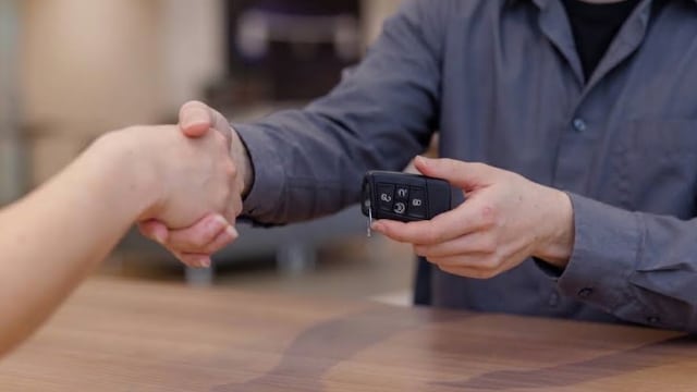 A person hands over car keys while shaking hands, symbolizing a successful car sale or rental.
